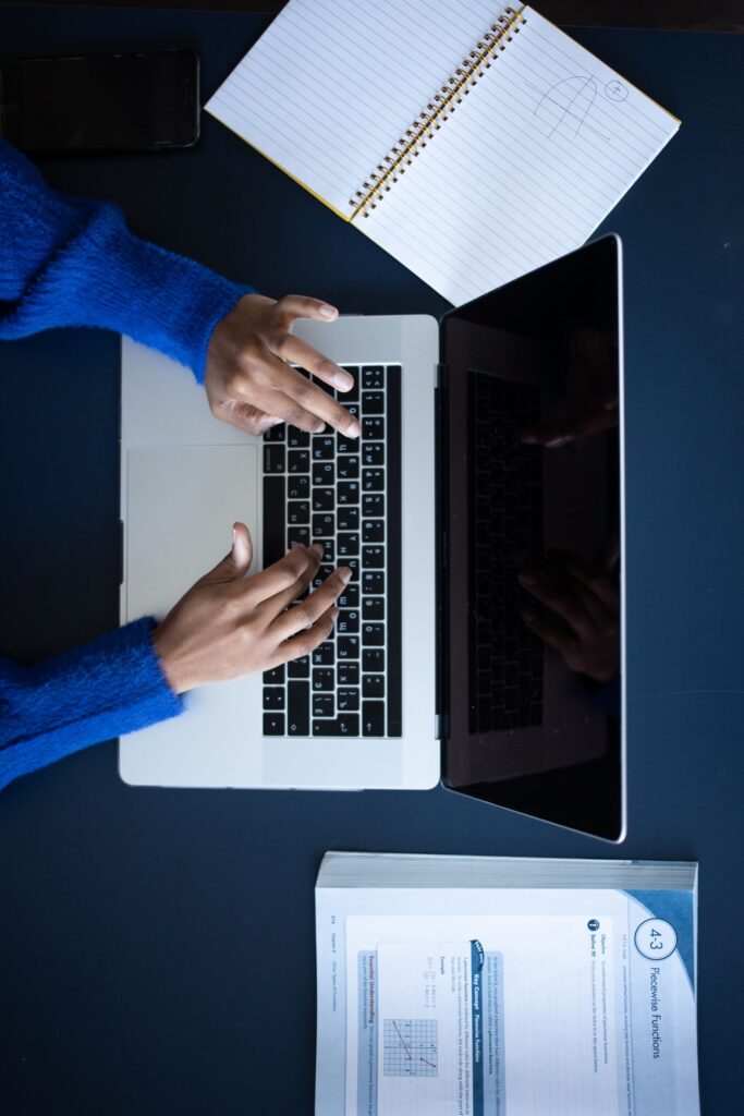 Overhead view of hands typing on a laptop next to a paper notebook